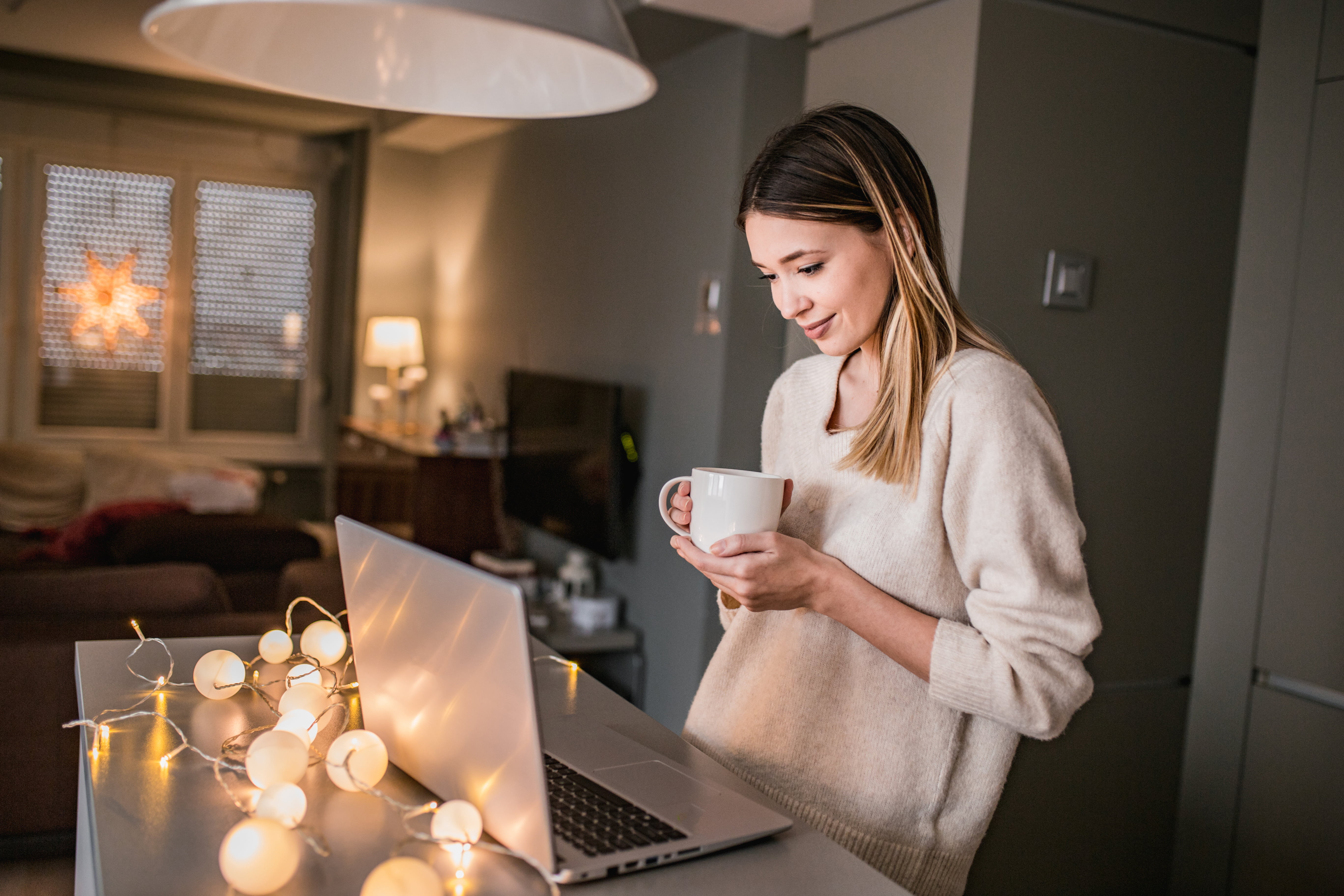 Kvinne i et nordisk hjem som sitter med laptop og kaffekopp i mykt vinterlys, med diskret julelys i bakgrunnen. Varm og rolig desemberstemning.