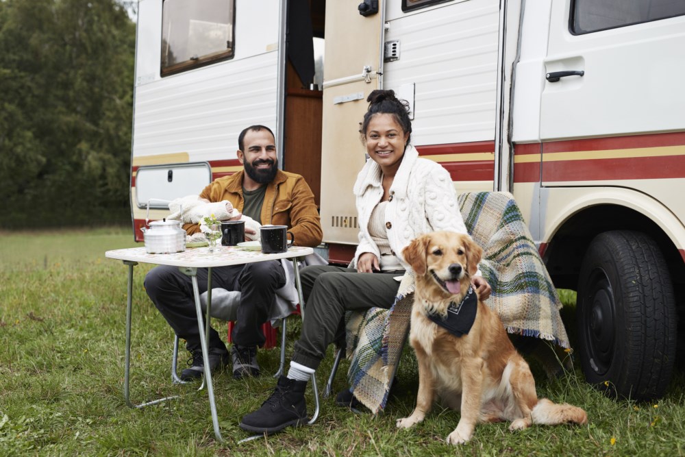 Et par smiler og drikker kaffe ved et bord utenfor deres bobil med en glad hund ved siden av dem, omringet av natur.